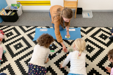 A woman kneels on the floor together with children and shows a game.